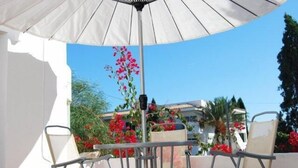 Un patio al aire libre con una mesa y sillas bajo un paraguas blanco, rodeado de flores rojas y vistas a edificios.