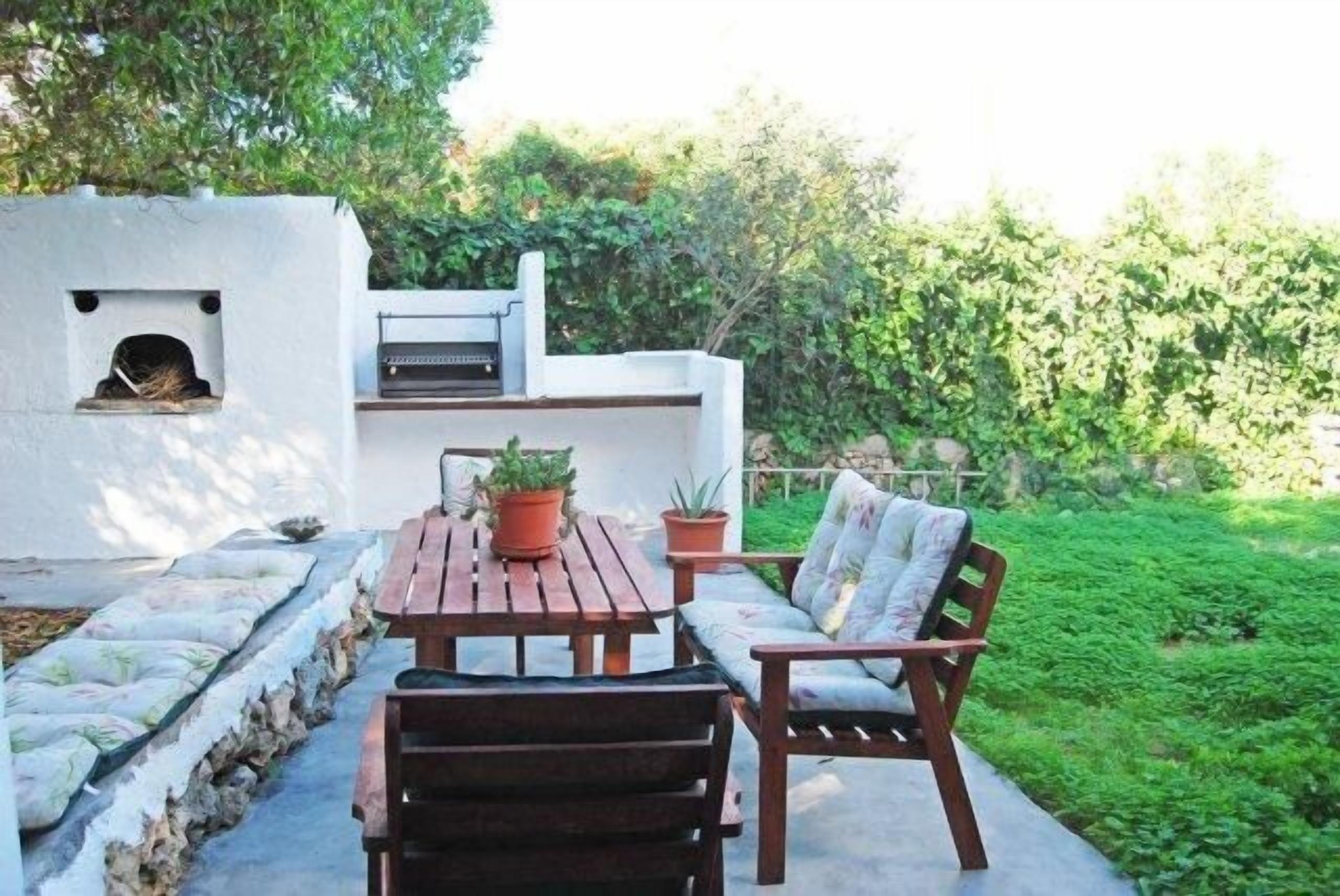 An outdoor patio with a wooden table and chairs, a barbecue grill, and a white wall with a small window.