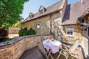 A stone building with a wooden deck, a table with a tablecloth, and a chair. - Stunning 16th Century Cotswold Manor House, Sleeps 12-14. (Chipping Norton)