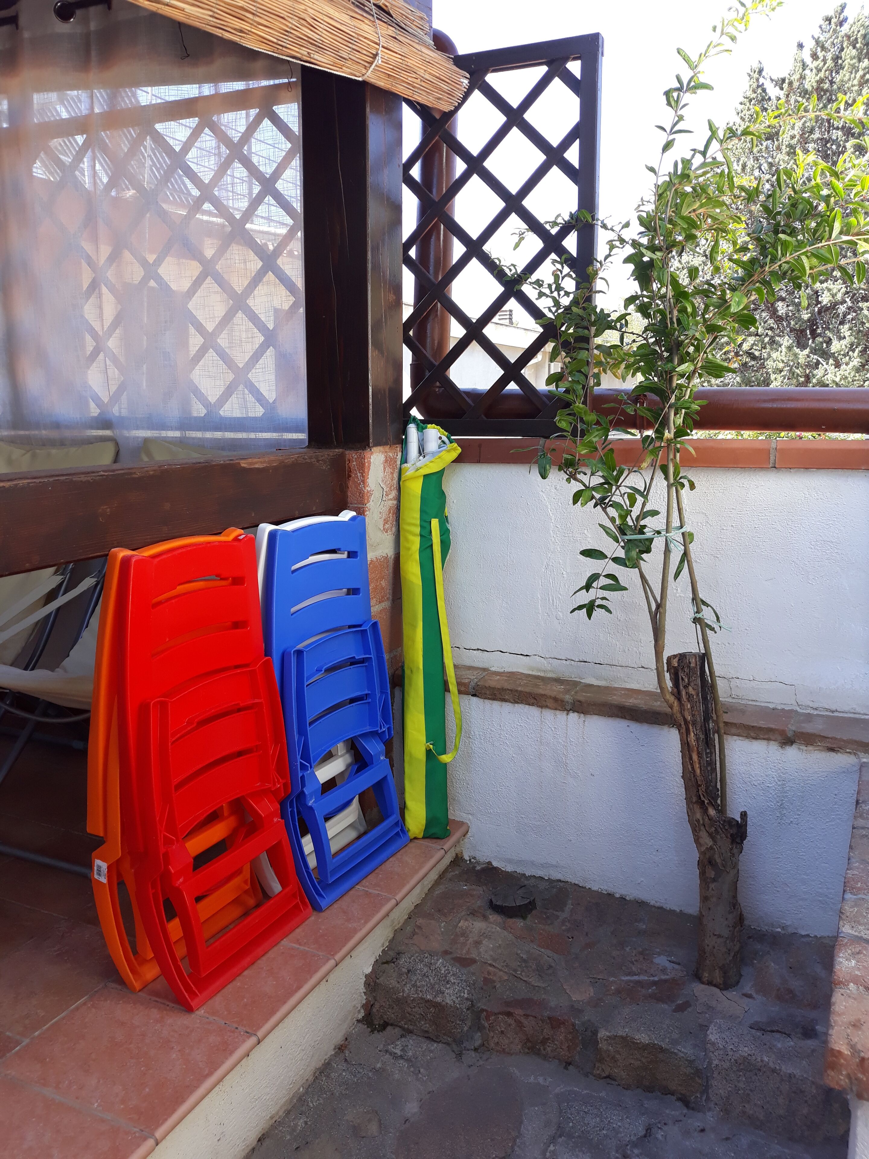 Un piccolo spazio all'aperto con una panca di legno, una pianta in vaso e sedie di plastica colorate.