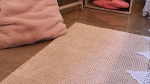 Una habitación con piso de madera, una manta rosa en una silla, una toalla blanca en un perchero, una pequeña mesita de noche con una lámpara y un mantel de encaje blanco.