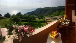 Un balcon avec une table dressée pour le dîner, un arrangement floral et une vue sur la mer et les collines.