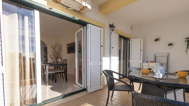 An open sliding glass door leading to a dining area with a table and chairs, a kitchen area with cabinets and a sink, and a small balcony with a table and chairs. - Casa Mateus (Alvor)