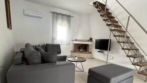 A modern living room with a grey sofa, a small round table, a TV, and a wooden staircase. - Beautiful villa with private pool & garden and stunning views of Navarone Bay. (Lindos)