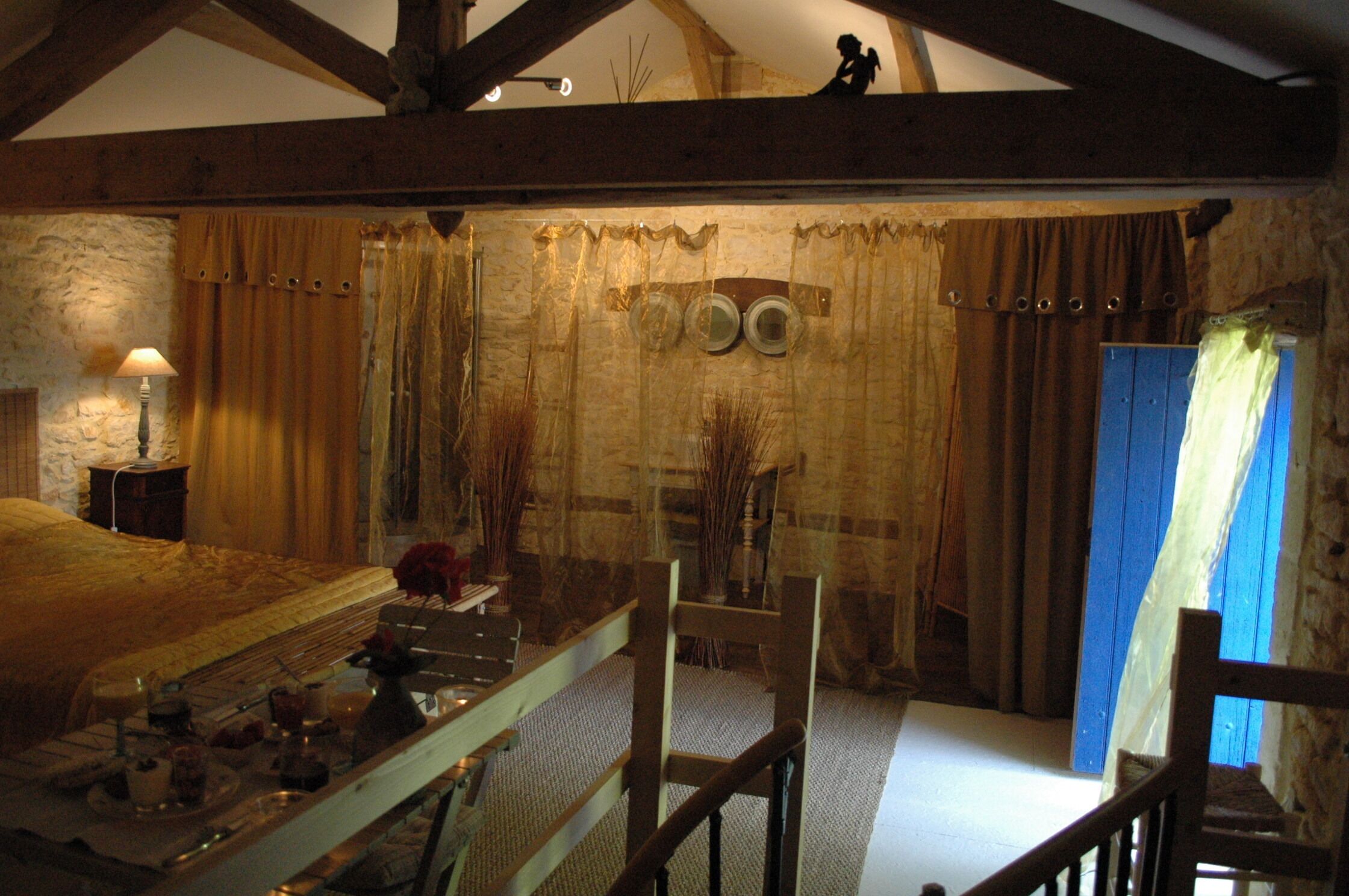 Une pièce rustique avec un mur en pierre, des poutres en bois, un lit doté d’une tête de lit en bois, une petite table avec une lampe et une porte bleue.