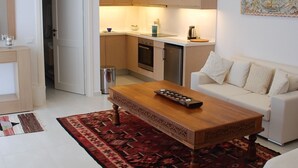 A modern living room with a wooden coffee table, a red patterned rug, a white sofa, and a kitchen area with wooden cabinets and a stove.
