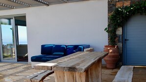 An outdoor seating area with a wooden bench and a sofa, a large terracotta pot, and a view of the sea through a glass door.
