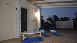 An outdoor patio area with a white table and blue chairs, a door, and a covered section with a view of trees. - Vendicari time, just steps away from the calamus beaches and vendors (NOTO)