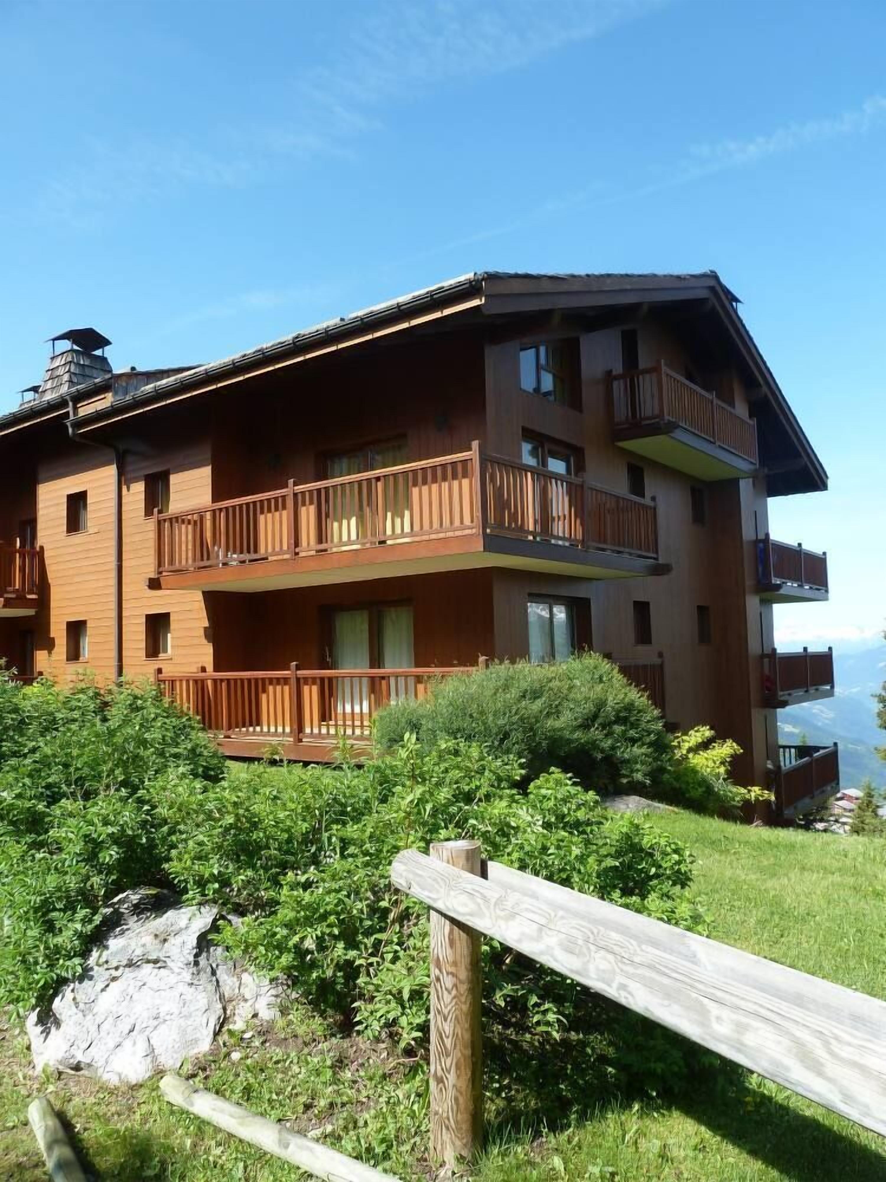 A multi-story wooden building with balconies, surrounded by greenery and a clear sky.