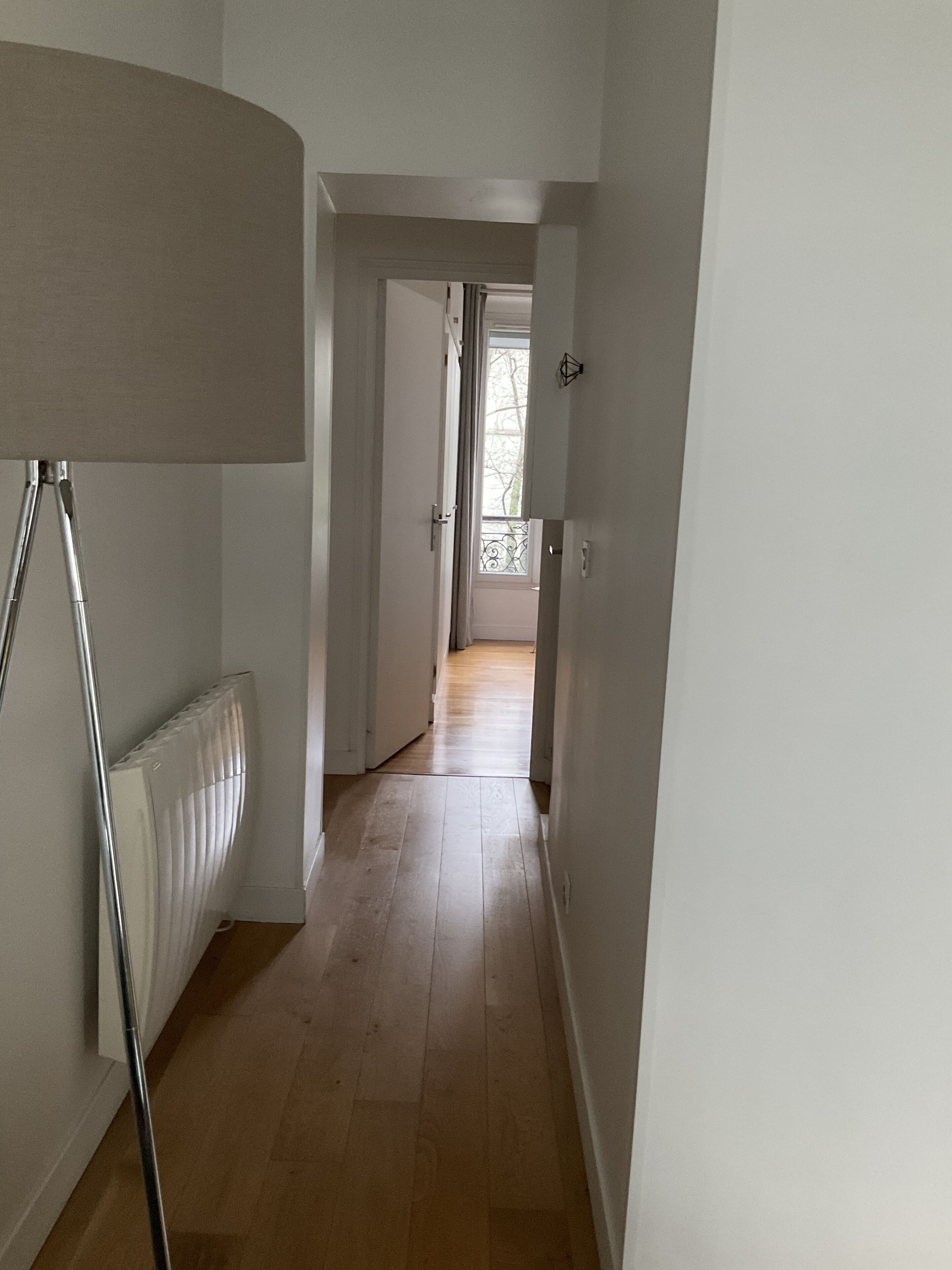 Un couloir avec un radiateur, une lampe sur pied et une vue sur une porte ouverte donnant sur un balcon.