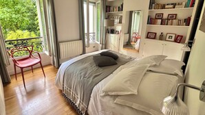 A bedroom with a bed, a chair, a bookshelf, and a view of greenery outside. - Charm and garden view, in the center of Paris, near Montparnasse (Paris)