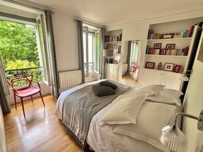 A bedroom with a bed, a chair, a bookshelf, and a view of greenery outside.