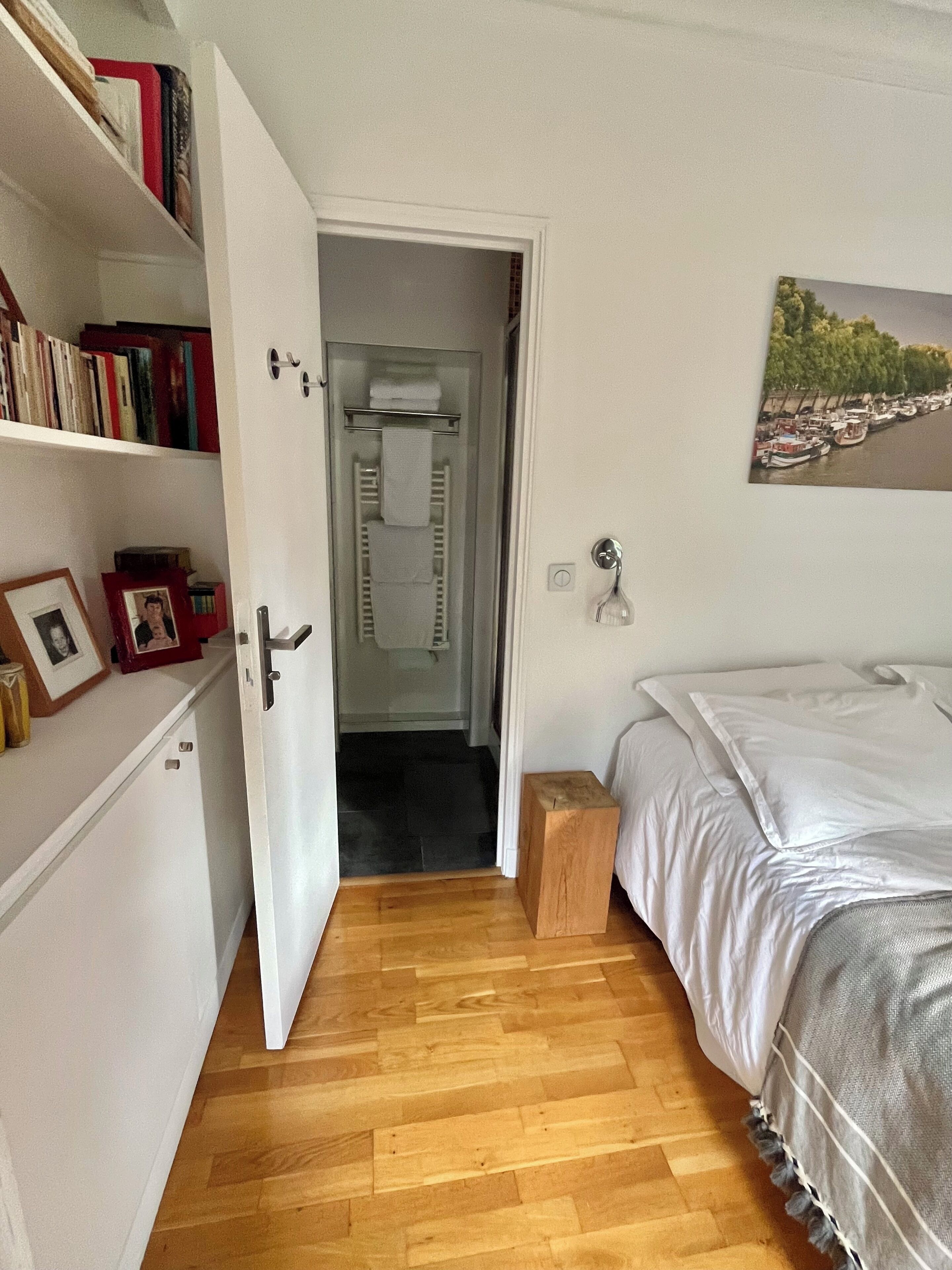 Une chambre avec un plancher en bois, un lit avec une literie blanche, une table de chevet, une bibliothèque et une porte donnant sur une salle de bain.
