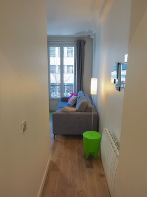 A small living room with a sofa, a green ottoman, a radiator, and a window with a view of buildings. - Charm and garden view, in the center of Paris, near Montparnasse (Paris)