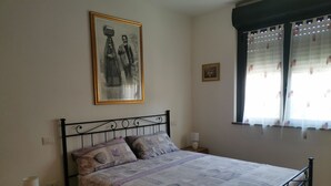 A bedroom with a bed, a nightstand, a drawer unit, a framed picture on the wall, and a window with curtains. - Apartment 2 km from the sea, 200 meters from the historic center of bosa (Bosa, Oristano)