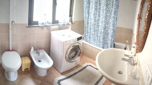 A bathroom with a toilet, bidet, washing machine, sink, and a window with a shower curtain. - Apartment 2 km from the sea, 200 meters from the historic center of bosa (Bosa, Oristano)