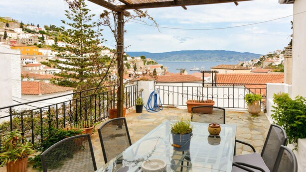 Un balcon avec une table en verre, des chaises, des plantes en pot et une vue sur une ville côtière.