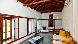 Une pièce avec un plafond en bois, un lit recouvert d’une courtepointe bleue et blanche, une chaise en bois, une petite table recouverte d’une nappe bleue et blanche, et une fenêtre donnant sur un paysage verdoyant.