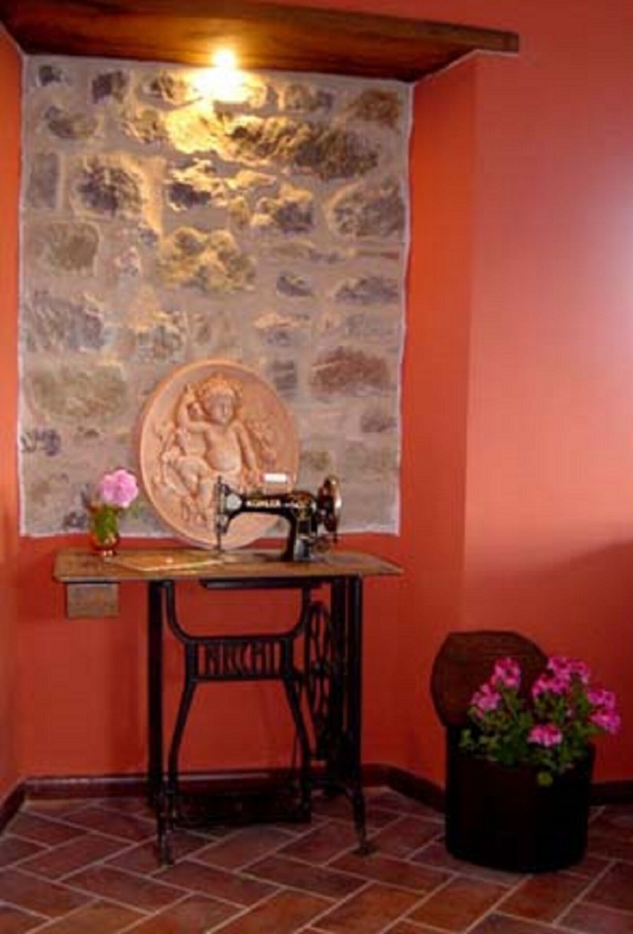 A room with a sewing machine on a wooden table, a tapestry on the wall, and a potted plant with flowers.