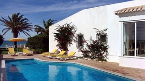 Une piscine avec des chaises longues jaunes, un palmier et un bâtiment blanc doté d’un toit en tuiles.