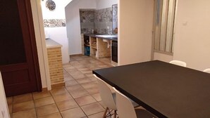 Un coin repas avec une table noire et des chaises blanches, une cuisine avec un mur recouvert de carreaux, et une horloge au mur.
