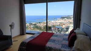 Camera d'albergo con una grande finestra che si affaccia su una città costiera, un letto con coperta rossa, un tavolino con sedie e una ventola da terra.