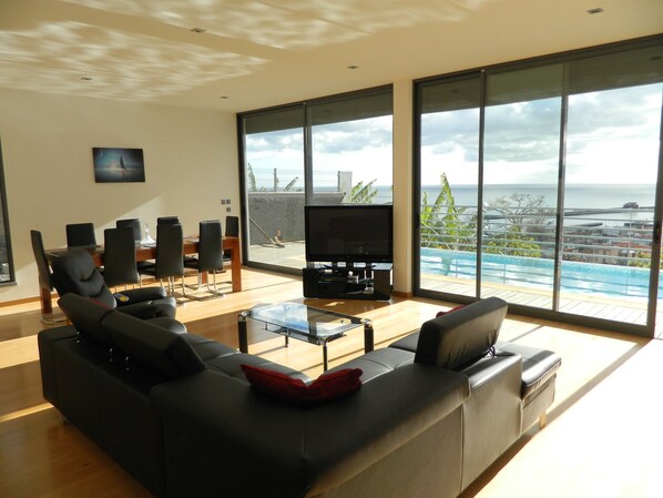 A modern living room with a large TV, comfortable seating, and a view of a pool and cityscape.