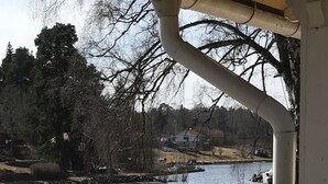 Une terrasse en bois avec des bancs, donnant sur une rivière paisible, entourée d’arbres et d’une maison située au loin.