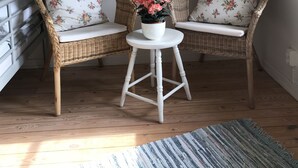Deux fauteuils en osier ornés de coussins à motifs floraux, une petite table blanche sur laquelle est posée une plante en pot, et quatre tableaux encadrés sur le thème de la marine, accrochés au mur.