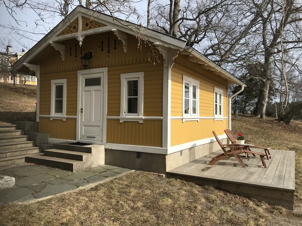 A small yellow cabin with a porch, steps leading to the door, and a wooden deck with chairs. - Cottage with sunny wooden deck by water. Rowing boat included. (Saltsjö-Boo)
