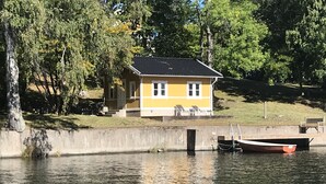 Une maison jaune avec un quai au bord d’un lac paisible, entourée d’arbres.