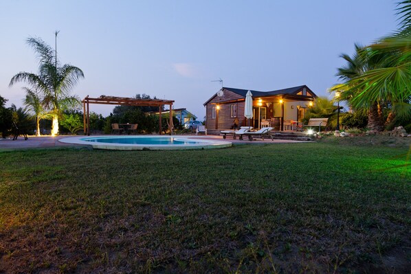Zona de piscina con sillas de descanso, una pérgola y una cabaña de madera con iluminación exterior.