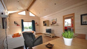 A cozy cabin interior with a wooden ceiling, a sofa, a dining table, a TV, and a potted plant.