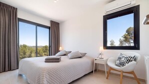 A modern bedroom with a large bed, a chair, a nightstand, and a window with a view of trees. - Luxury vacation villa in the hills of Sitges with breathtaking views (Olivella)