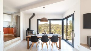 Une cuisine moderne dotée d’une cuisinière intégrée, un coin repas avec une grande table et des chaises, et une vue sur un paysage vallonné grâce à de grandes fenêtres.