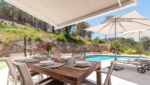 Un espace repas extérieur avec une table en bois dressée pour quatre personnes, entourée de chaises blanches, sous un grand parasol. On aperçoit une piscine à l’arrière-plan.