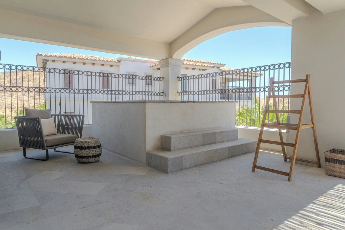 A balcony with a wrought iron railing, a wooden ladder, a woven ottoman, and a cushioned chair.