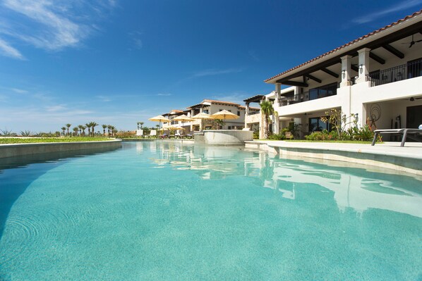 A clear swimming pool with a view of a resort building with balconies and palm trees in the background.