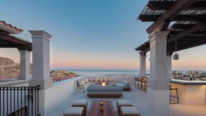 An outdoor seating area with a view of the sea, featuring a long wooden table, cushioned seating, and a pergola structure. - Live Aqua Private Residences Los Cabos (Cabo San Lucas)
