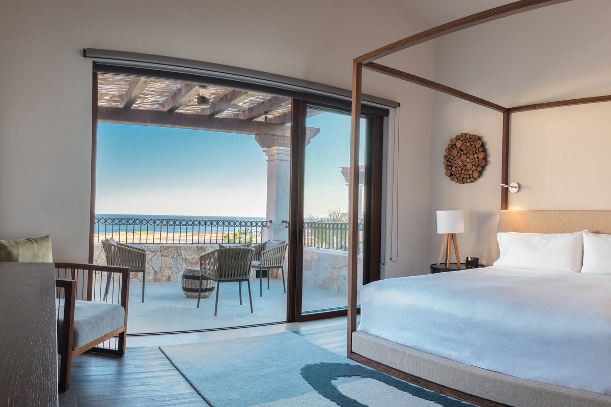 Een hotelkamer met een hemelbed, een balkon met zitplaatsen buiten en uitzicht op zee.