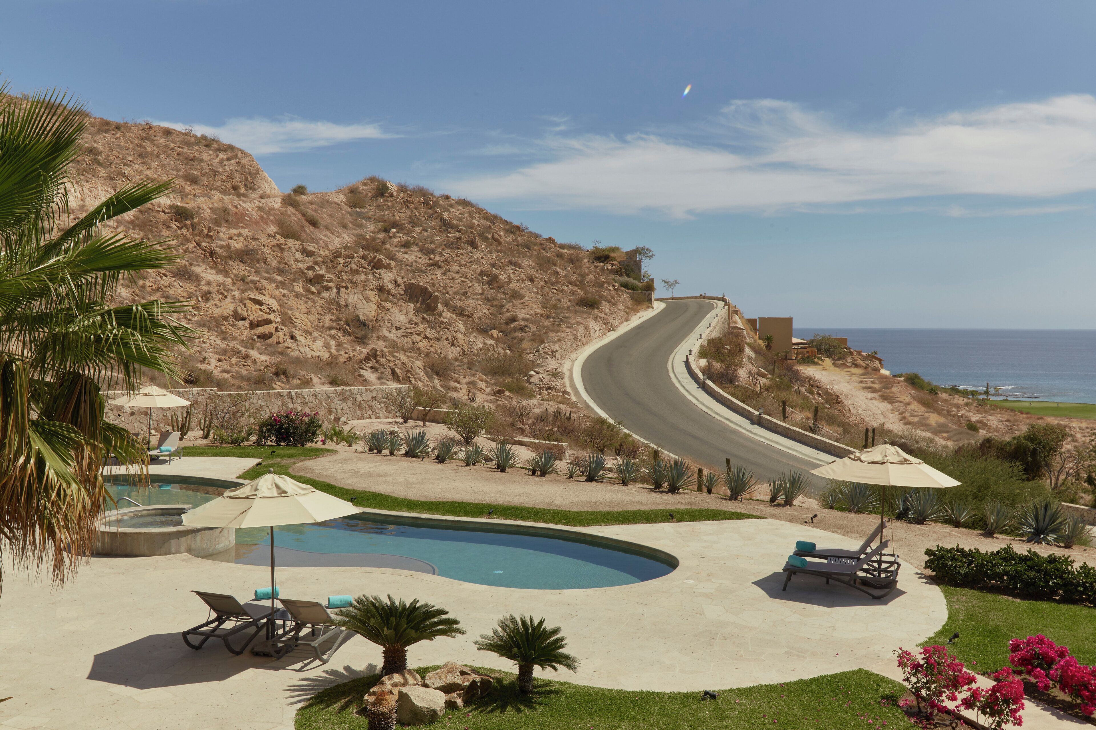Un espace piscine avec des chaises longues et des parasols, un chemin sinueux et une vue sur l’océan.