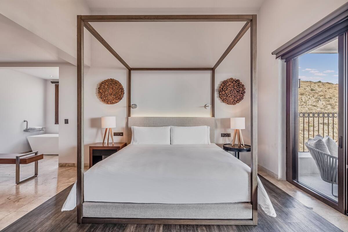 Une chambre moderne avec un grand lit, des tables de chevet avec des lampes, une vue sur le désert par une porte coulissante, et une salle de bain avec une baignoire et un lavabo.