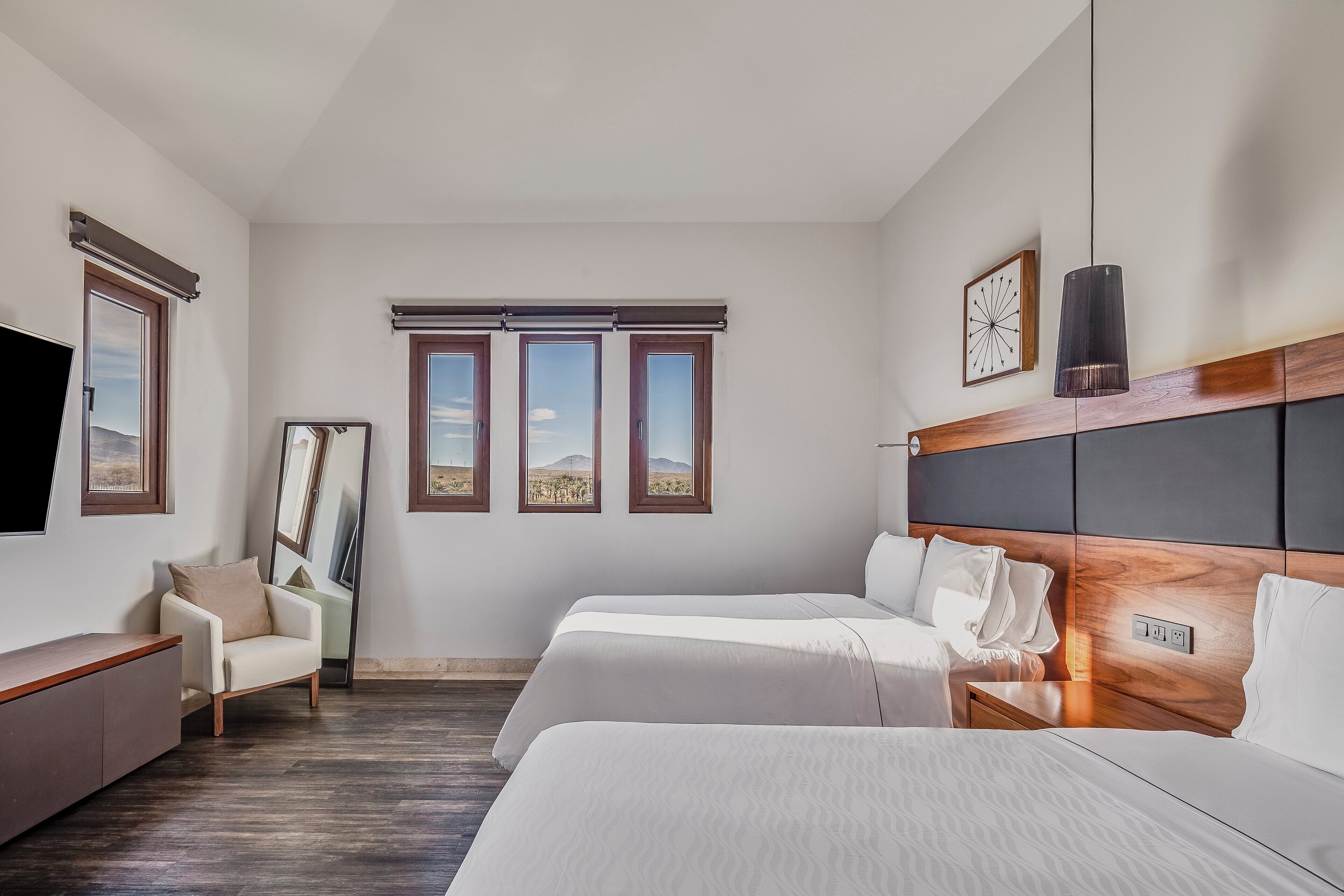 Ein Hotelzimmer mit zwei Betten, einem Schlafsofa, einem Fernseher und einem großen Fenster mit Blick auf eine Wüste.