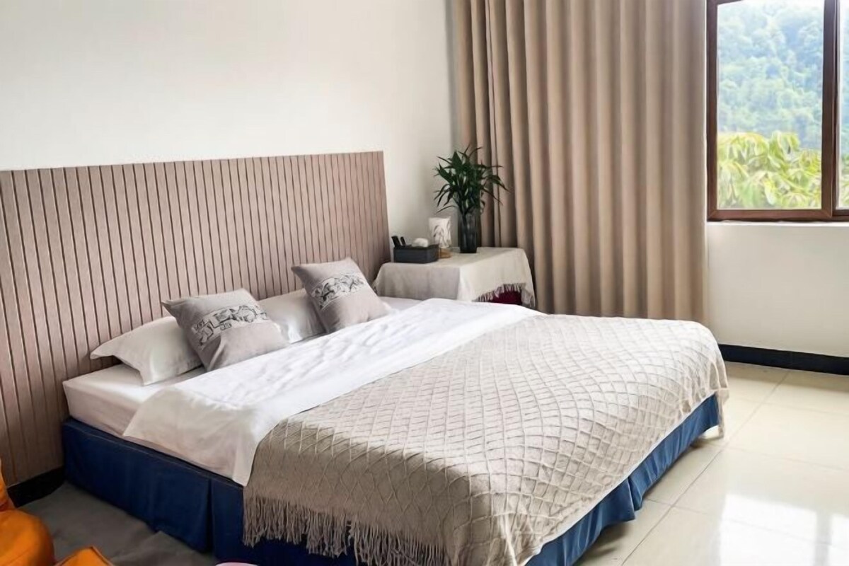 A neatly made bed with a blue skirt, a headboard with vertical lines, a bedside table with a plant, and a window with a view of greenery.