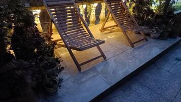 Two wooden lounge chairs with metal frames, positioned on a tiled patio with potted plants and a stone wall in the background.