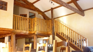 A spacious room with a high ceiling, wooden beams, a balcony with a railing, a staircase, and a dining area with chairs and a table. - Country Cottage / Gite - Usso, en Forez (Usson-En-Forez)