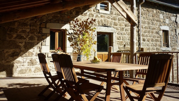 An outdoor wooden table and chairs set against a stone building with windows and a wooden beam structure overhead. - Country Cottage / Gite - Usso, en Forez (Usson-En-Forez)