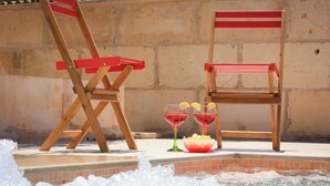Deux chaises en bois et deux verres d’une boisson rouge, près d’une piscine.