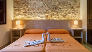 Une chambre d’hôtel avec un mur en pierre à la texture particulière, deux lampes de chevet, une couvre-lit orange et deux serviettes en forme de cygne posées sur le lit.