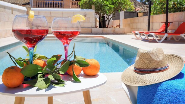 Une scène au bord de la piscine avec deux verres remplis d’une boisson rouge, des oranges et un chapeau de paille sur une table.